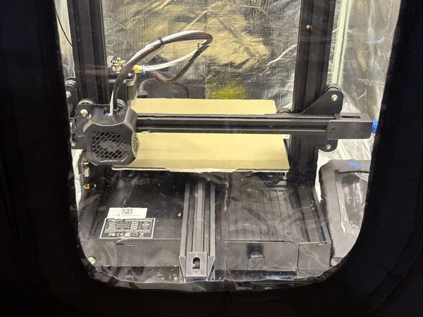 Looking into a 3d printer enclosure through a plastic window. Inside, a Ender 3 v2 is well-lit.