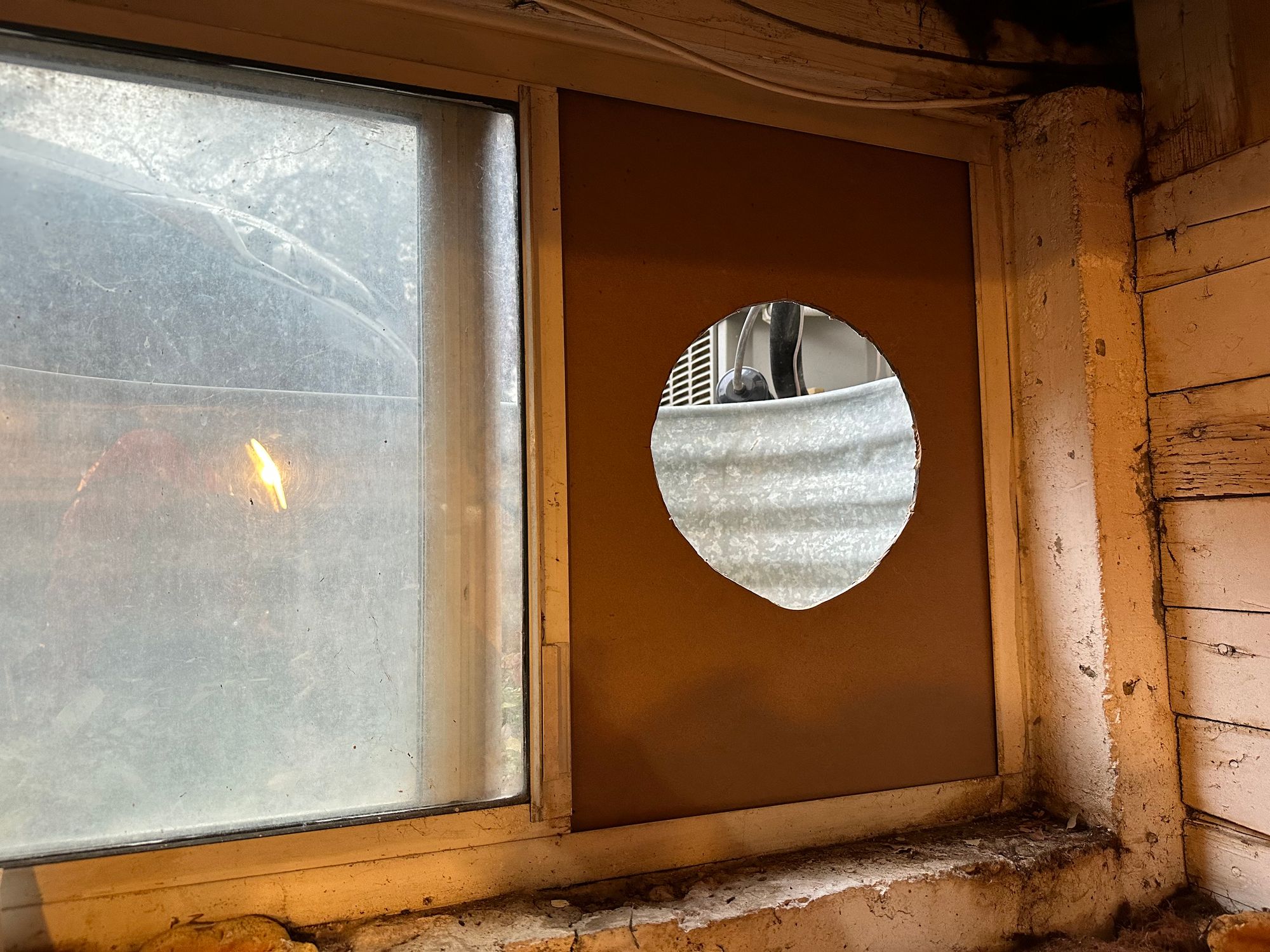 The window well in my basement workshop, with the window open and a piece of tileboard inserted. The tileboard has a rough circular hole cut into it for venting air from the paint booth.