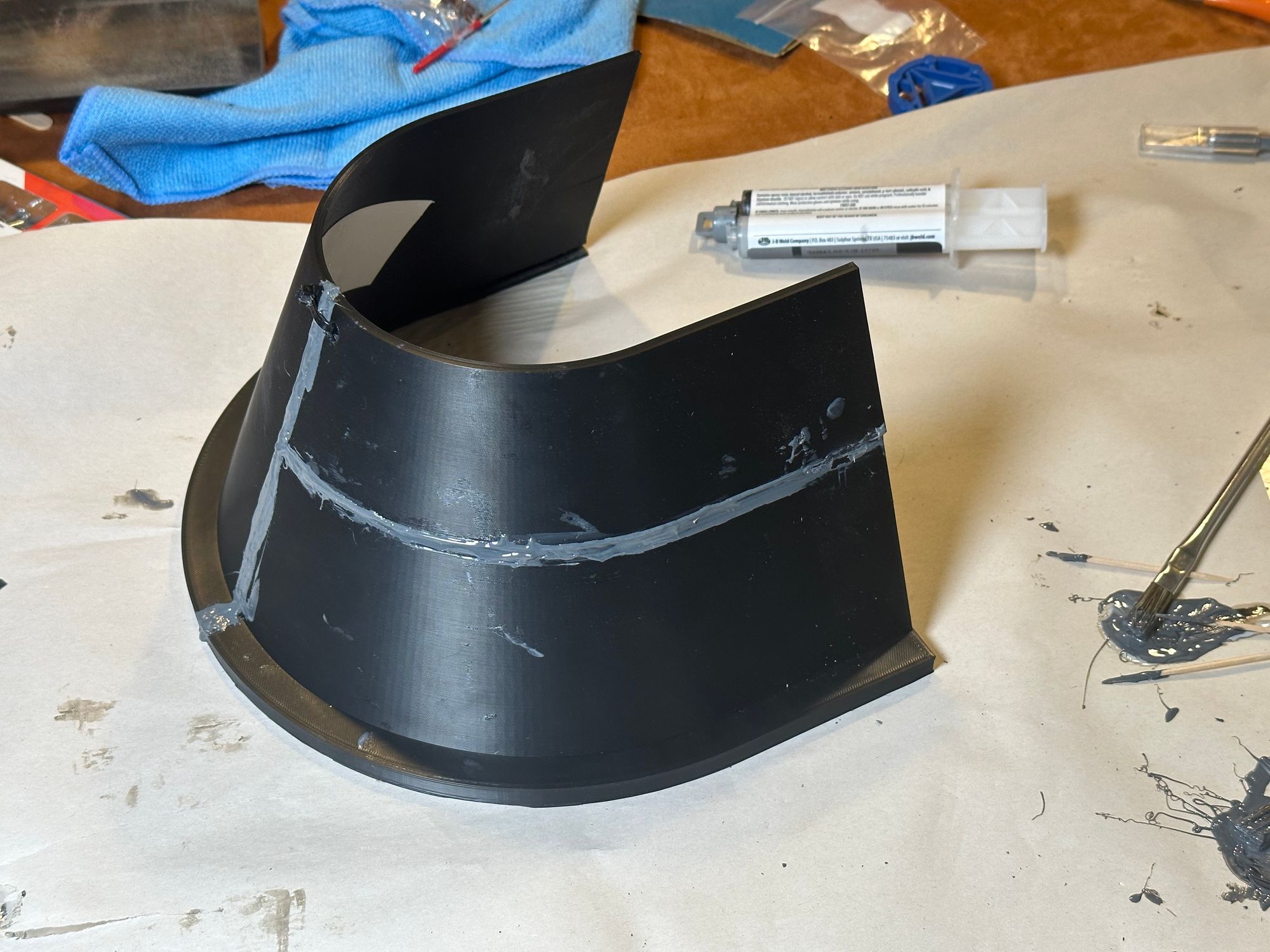 Both pieces of the hood, epoxied together. The gray epoxy is very noticeable, both down the center seam, and along a horizontal seam where one piece split. A zip tie secures the end pieces of the hood together, and some white Gorilla tape is visible on the underside. Leftover epoxy and disposable tools sit nearby.