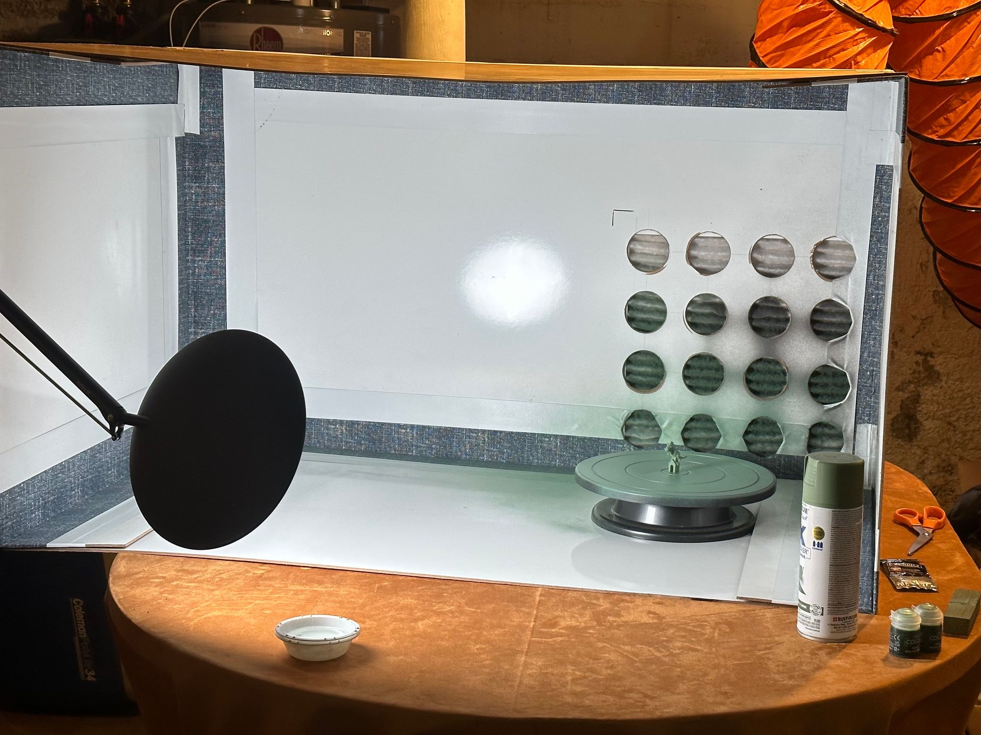 The paint booth, set up on its side, after painting a small piece. Inside the booth is a painting turntable and the piece, and paint supplies sit nearby, illuminated by a lamp.