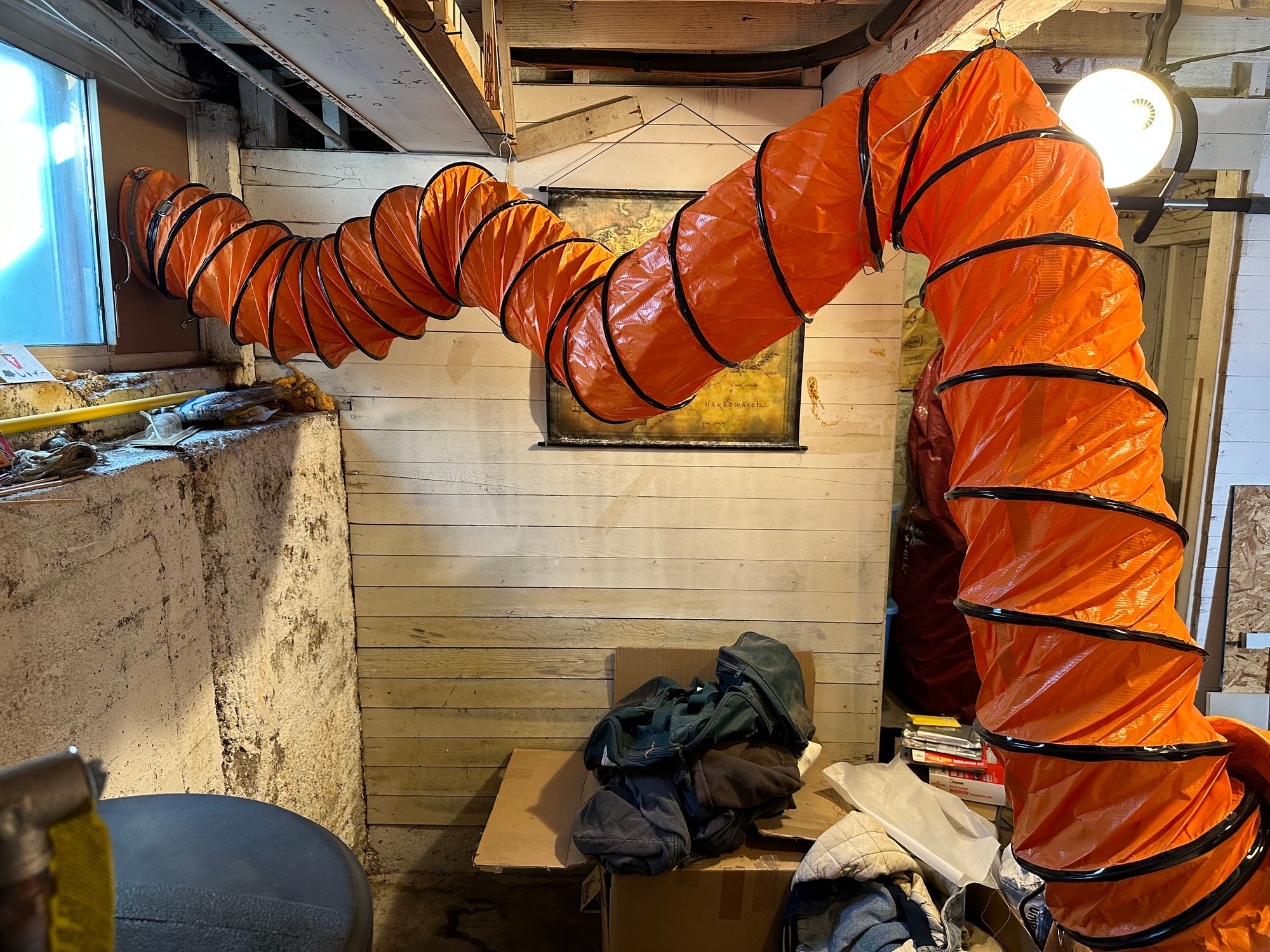 An orange ducting hose, which is suspended in two places between the blower and the window. A messy basement is seen in the background.